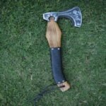 Hand-forged Viking axe with a braided leather grip, featuring a carbon steel blade and a wooden handle, displayed on a grassy background